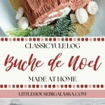 A homemade Bûche de Noël, decorated with meringue mushrooms and greenery, is displayed on a white platter with a sprinkle of brown sugar.