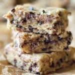 A stack of three chocolate chip cookie bars, reminiscent of banana bars, on parchment paper. The bars have a golden-brown crust and are filled with visible chocolate chips.
