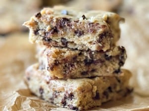 A stack of three chocolate chip cookie bars, reminiscent of banana bars, on parchment paper. The bars have a golden-brown crust and are filled with visible chocolate chips.