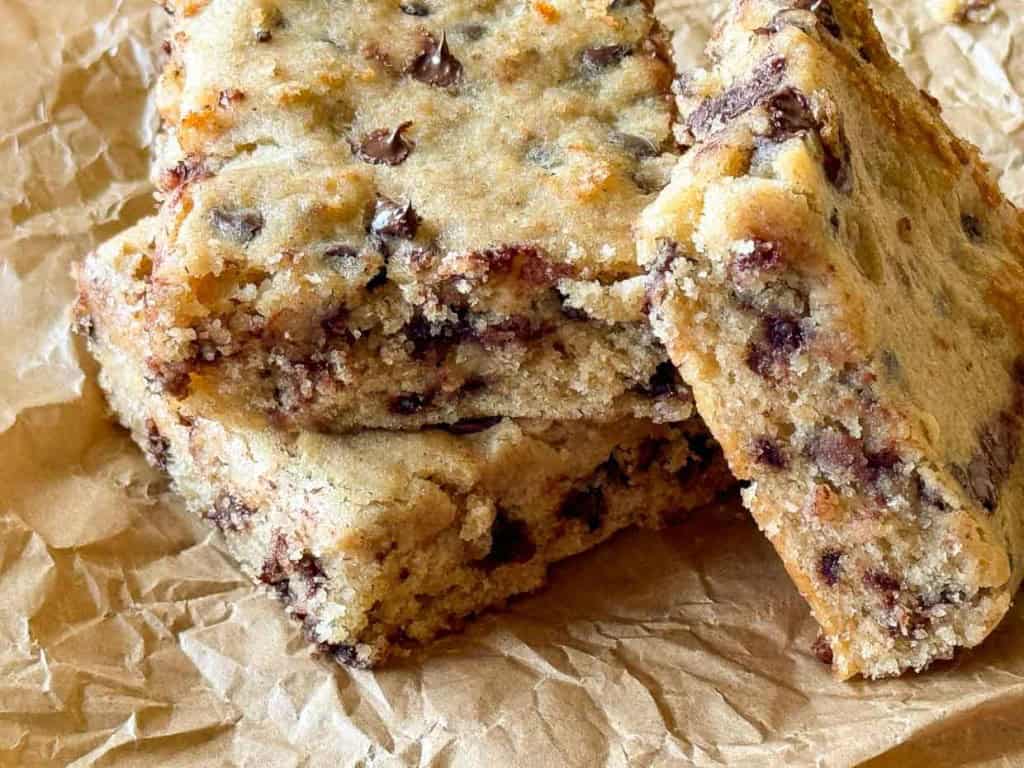 Close-up of three chocolate chip cookie bars stacked on a piece of crumpled parchment paper. The bars, reminiscent of Banana Bars, have a golden-brown crust and visible chocolate chips throughout.