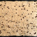 A pan of raw chocolate chip cookie dough spread evenly with some chocolate chips visible on the surface, reminiscent of a batch of decadent Banana Bars.