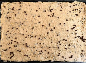A pan of raw chocolate chip cookie dough spread evenly with some chocolate chips visible on the surface, reminiscent of a batch of decadent Banana Bars.
