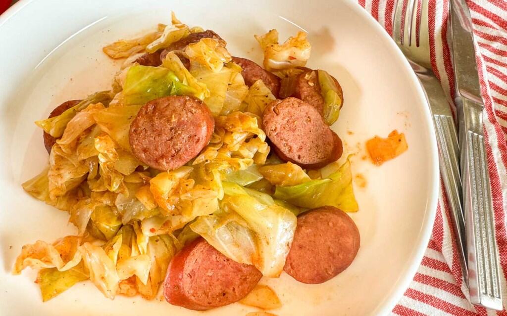 A plate of smothered cabbage with smoked sausage sits invitingly on a white plate, accompanied by a red and white striped cloth and gleaming silverware.