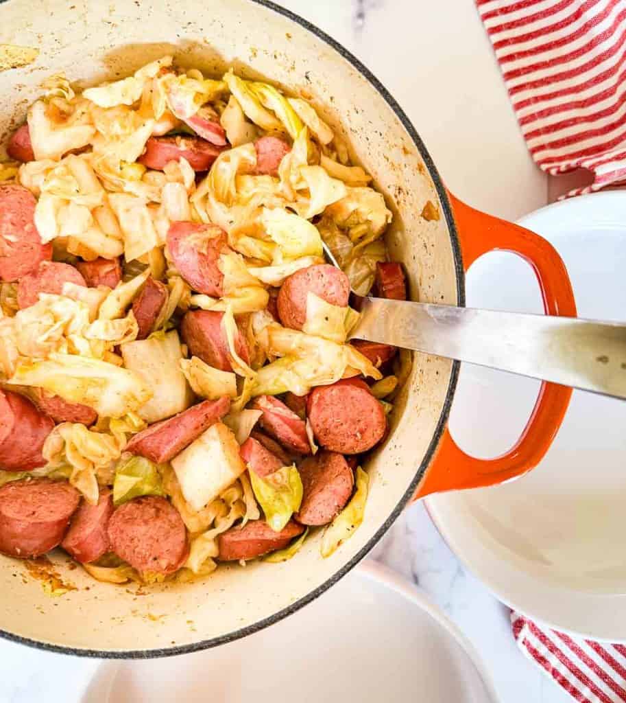 In an orange Dutch oven, the smothered cabbage with smoked sausage is cooked to perfection. A metal serving spoon rests ready beside a red and white striped cloth, promising a deliciously hearty meal.