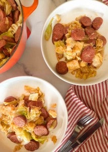 Two plates of smothered cabbage with smoked sausage sit beside a red-striped cloth, while a pot brimming with more of the savory dish peeks into view.