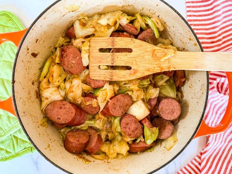 A pot with fried cabbage and sliced sausage, seasoned to perfection. A wooden spatula rests inside. Nearby are a red-striped cloth and a green oven mitt, ready for the culinary odyssey of flavors in smothered cabbage with smoked sausage.