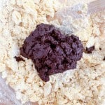 A mound of chocolate dough, destined for Texas Sheet Cake Cookies, sits atop a pile of pale, crumbly dough, with a wooden spoon partially mixing the ingredients.