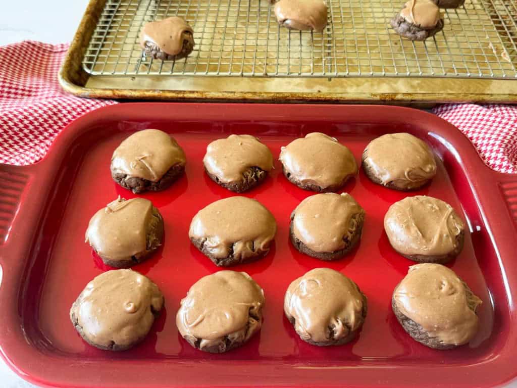 Texas Sheet Cake Cookies, chocolate-frosted to perfection, are arranged in a red baking dish, with more cookies cooling on a wire rack over a sheet pan in the background.
