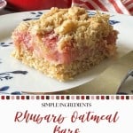 A single rhubarb oatmeal bar sits on a plate, topped with crumbly oats—the perfect example of delicious homemade Rhubarb Bars. A baking dish and a striped cloth are in the background.