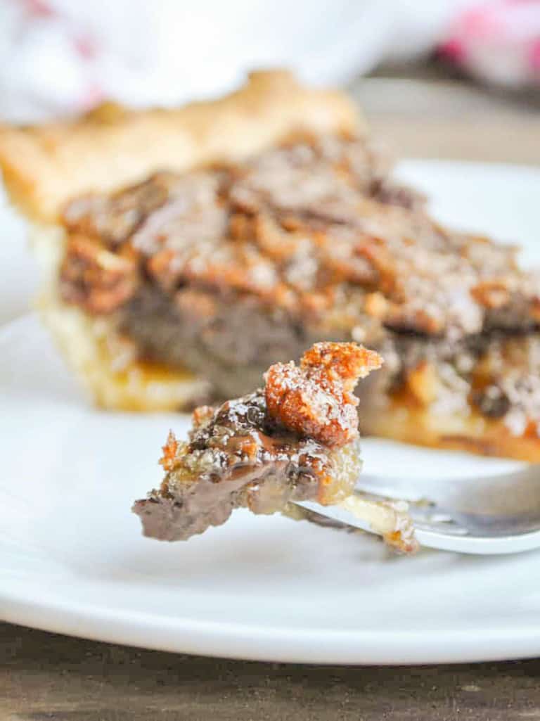 A fork holding a bite of chocolate pecan pie, with a slice of the pie from our recipe in the background.