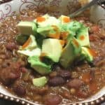 A bowl of vegan lentil topped with slices of avocado and chili sauce.