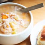 A tighter shot of the fresh homemade bean soup recipe. This picture has a bowl of soup, with a spoon tucked in it, and small part of the bread roll on a plate is showing.