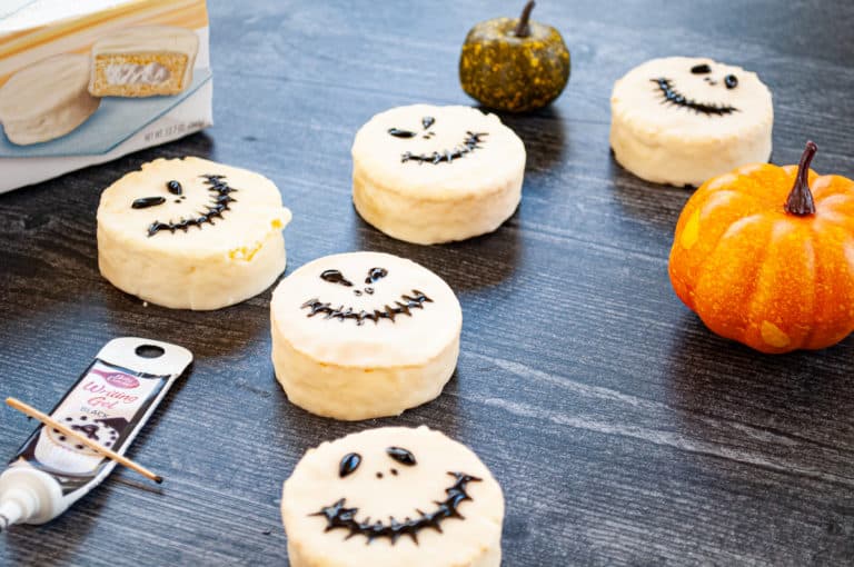 Jack Skellington Halloween treats, decorated with black writing gel, on a wooden surface next to a pumpkin and a box of White Fudge Ding Dongs.
