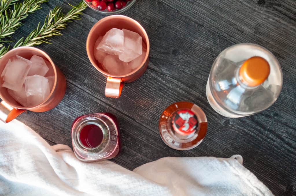 Two copper mugs with ice, a bottle of liquid for a Cranberry Moscow Mule, a jar of red sauce, and a bowl of berries on a dark wooden table.