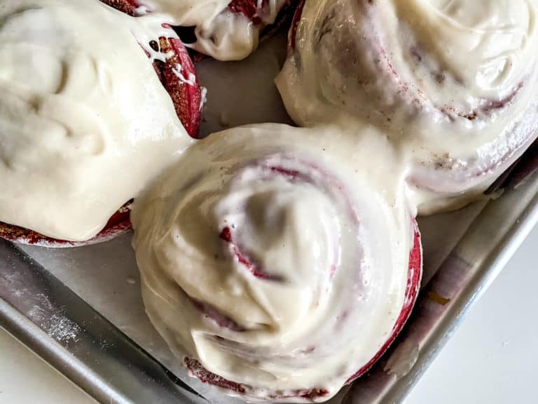 Freshly baked red velvet cinnamon rolls with cream cheese frosting on a baking tray.
