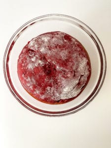 A roll of red velvet dough, doubled in size after rising, sits in a glass bowl.