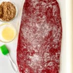 A red velvet dough rolled out on a surface with flour, a bowl of brown sugar, melted butter in a bowl, a pastry brush, and a rolling pin nearby.