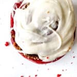 A top view of Red Velvet Cinnamon Rolls with a creamy topping on a white surface with crumbs scattered around.
