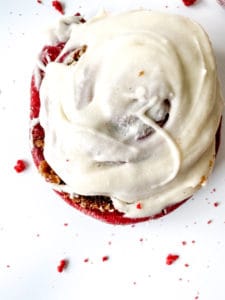 A top view of Red Velvet Cinnamon Rolls with a creamy topping on a white surface with crumbs scattered around.