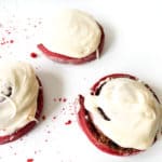 Red velvet cinnamon rolls with cream cheese frosting on a white surface, some with crumbs scattered around.