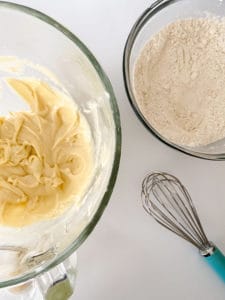Ingredients prepared for baking cream cheese sugar cookies with a bowl of creamed butter and sugar next to a bowl of flour, accompanied by a whisk.