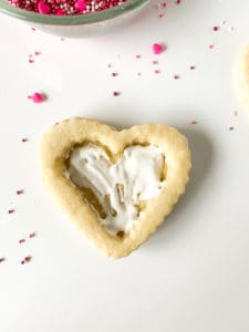 A Red Velvet heart-shaped cookie with white icing on a white surface, surrounded by colorful sprinkles.