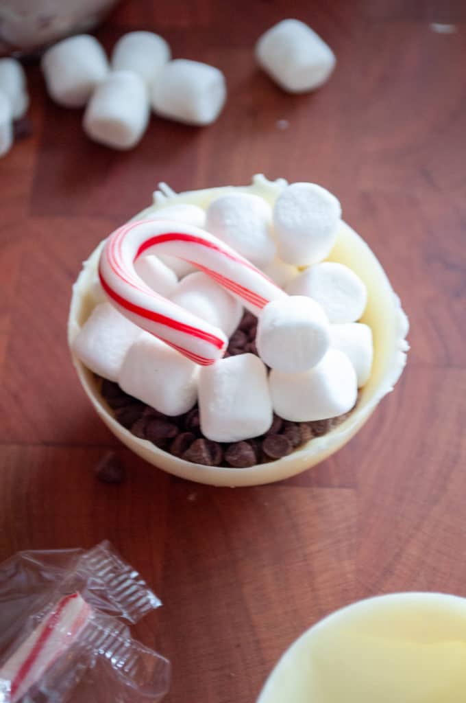 A bowl of chocolate chips topped with marshmallows, a candy cane on a wooden surface, is reminiscent of Snowman Soup.