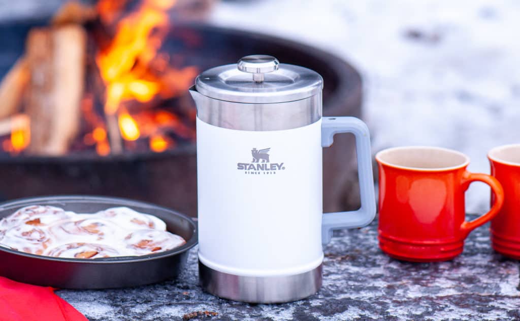 A Stanley Classic Hot French Coffee Press next to two mugs and a pan of cinnamon rolls on a snowy surface with a campfire in the background.