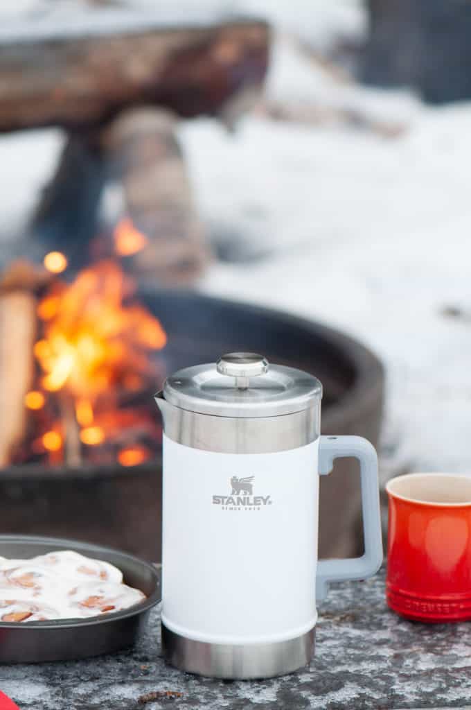 A Stanley Classic Stay Hot French Coffee Press, mug with coffee, and cinnamon buns on a wooden surface beside a campfire.