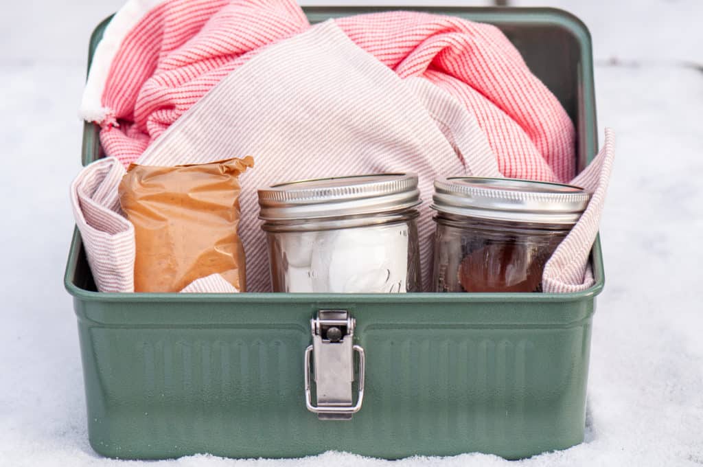 A green Stanley Brand metal box with jars and a pink-striped cloth on a snowy surface.