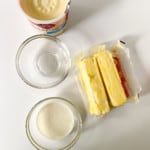 A tub of store-bought frosting, a stick of butter, cream, and a glass bowl are placed on a white surface.