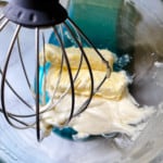 An electric mixer whisk attachment covered in semi-homemade buttercream, inside a glass mixing bowl.