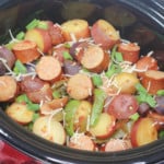 A slow cooker filled with a meal of sausage and potatoes, green beans, and garnished with herbs.