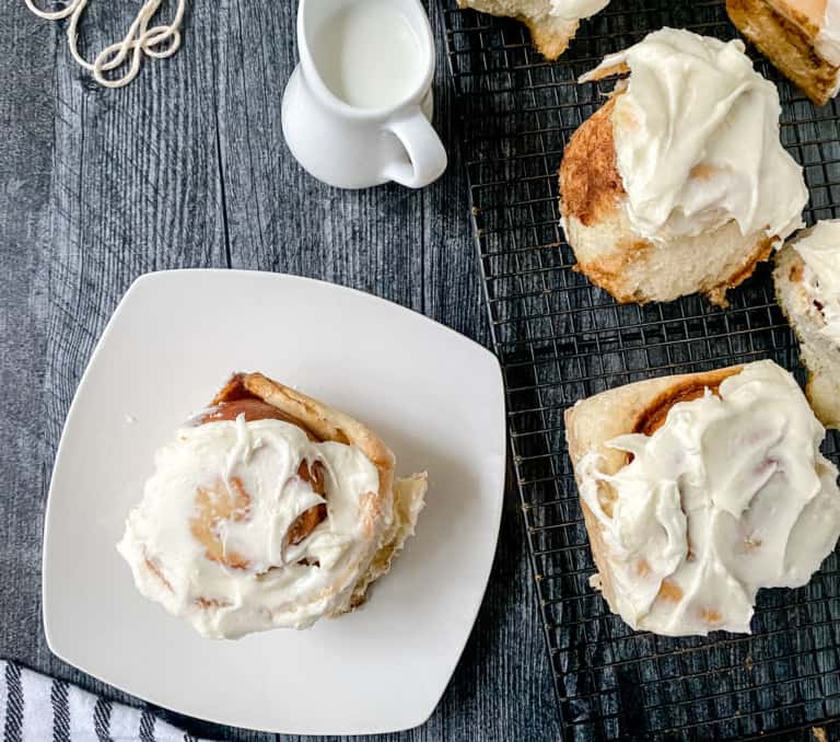 Freshly baked cinnamon rolls with creamy frosting on a wire rack and white plate, served with a side of milk.