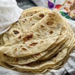 A stack of freshly made flatbreads with golden-brown spots, wrapped in a clean white cloth on a table.