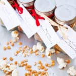 Glass jars with labels for popcorn seasoning mix, tied with red ribbons, beside loose kernels on a surface.