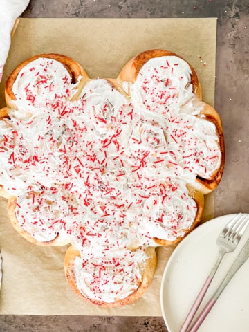 Large heart made of cinnamon rolls frosted with whtie frosting and sprinkled over with sprinkles.