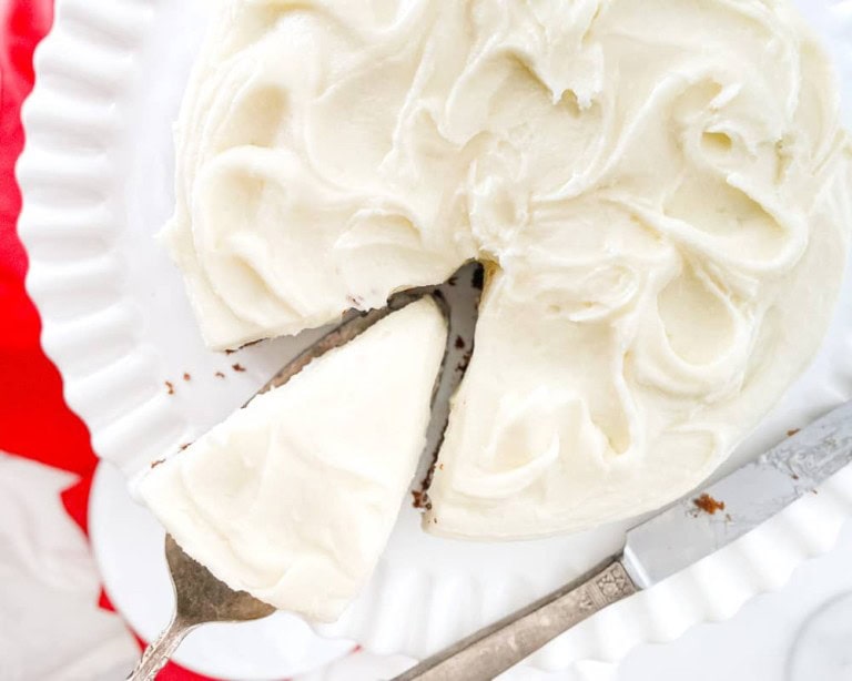 A slice of gingerbread cake with creamy frosting on a white plate, next to the rest of the cake.