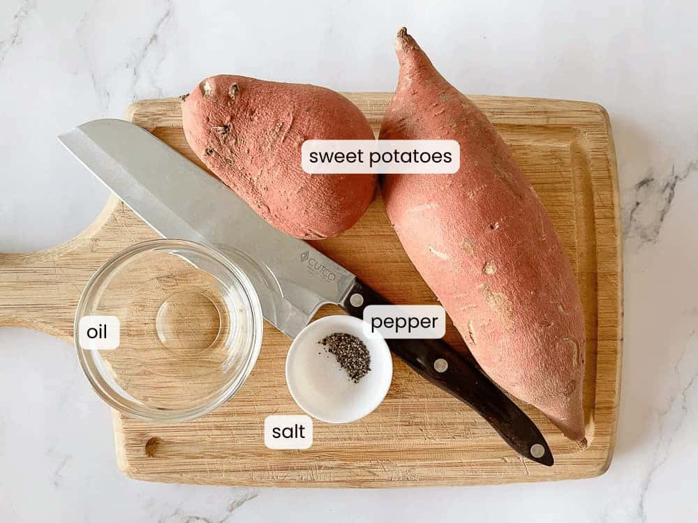 Two raw sweet potatoes, a bowl of oil, a small dish of salt and pepper, and two knives are arranged on a wooden cutting board—everything you need to prepare delicious AIR FRYER ROASTED SWEET POTATOES.