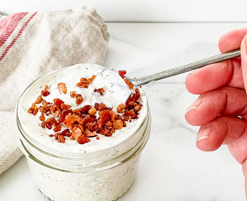 Ranch dressing with bacon bits on top with a spoon dipped into the jar.