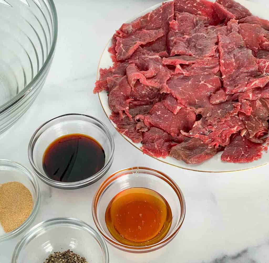 Thin slices of raw beef on a plate next to glass bowls containing soy sauce, honey, ground black pepper, and garlic powder on a white surface—perfect ingredients if you’re looking for how to make beef jerky or searching for an easy beef jerky recipe.