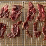 Several pieces of seasoned raw beef are arranged on a metal cooling rack, prepared for drying or making jerky—an essential step in any great beef jerky recipe and a perfect example of how to make beef jerky at home.