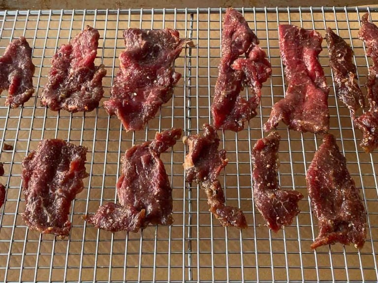 Several pieces of seasoned raw beef are arranged on a metal cooling rack, prepared for drying or making jerky—an essential step in any great beef jerky recipe and a perfect example of how to make beef jerky at home.