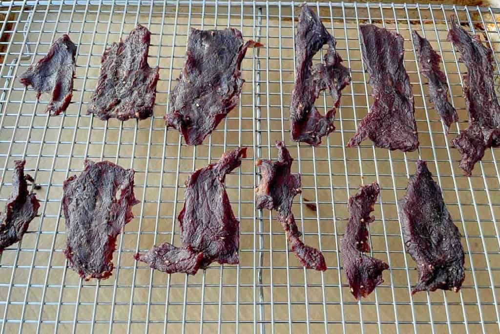 Thin strips of beef jerky, made following a classic beef jerky recipe, are arranged on a metal cooling rack placed over parchment paper.