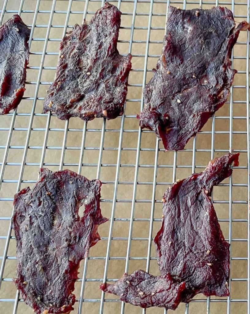 Four pieces of beef jerky are drying on a metal cooling rack, showcasing the results of a classic beef jerky recipe. The jerky pieces vary in shape and size, highlighting the hands-on process of how to make beef jerky at home. The grid rack is placed over a tan surface.