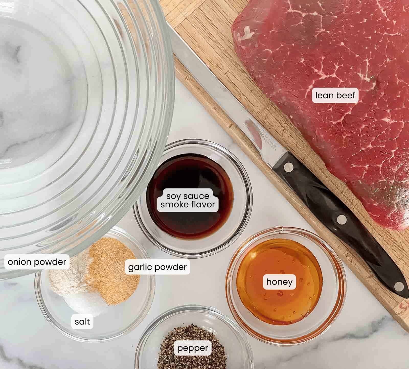 A glass bowl, raw lean beef ready for your favorite beef jerky recipe, a knife, and small bowls of onion powder, garlic powder, soy sauce with smoke flavor, honey, salt, and pepper are all arranged on a kitchen counter—everything you need for how to make beef jerky at home.