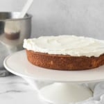 A frosted strawberry sponge cake on a white plate with a metal mixing bowl and a whisk in the background.