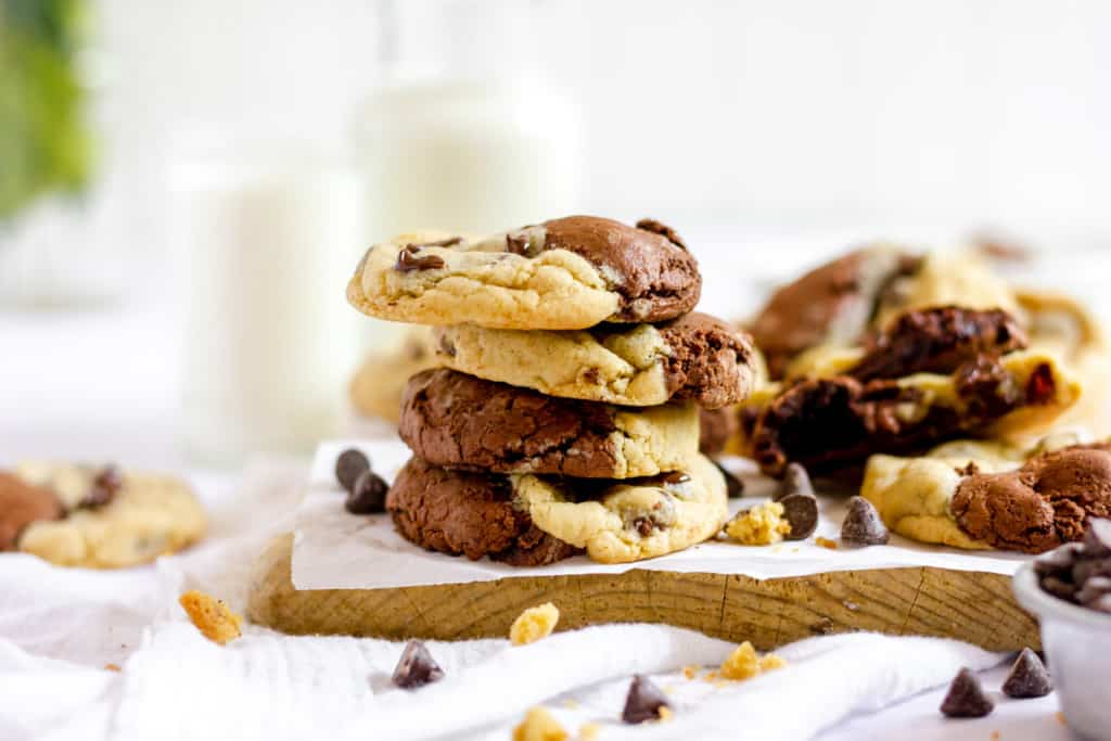 A stack of chocolate chip cookies, some resembling brookies with a mix of chocolate and vanilla dough, on a wooden board with milk bottles in the background. Chocolate chips are scattered around.