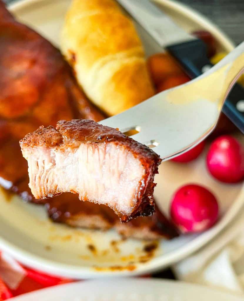 A fork holds a piece of cooked meat above a plate with barbecue ribs, an air fryer pork steak, a crescent roll, a knife, and several red grapes.
