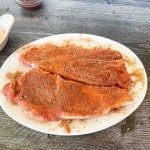 Two raw pork ribs covered in dry spice rub rest on a white oval plate, sitting on a wooden table&mdash;perfectly prepared for making delicious air fryer pork steak. A small dish and a bowl are partially visible nearby.